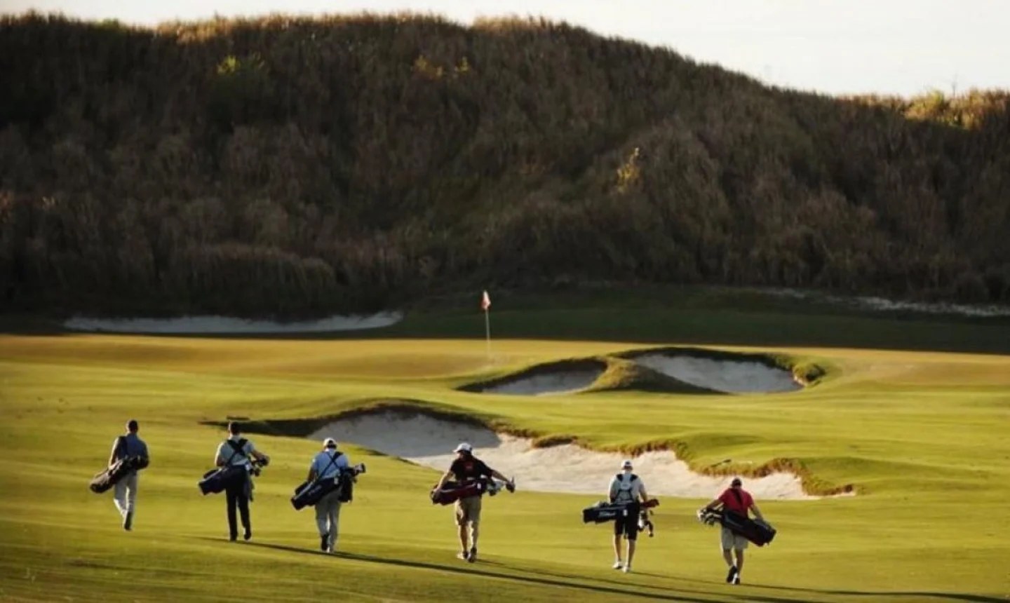 Six golfers carrying their bags down a sunlit fairway past deep bunkers